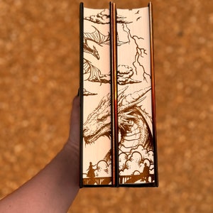 May include: Two hardback books with cream-coloured covers featuring a dragon illustration. The dragon is depicted flying through clouds with lightning bolts in the background. The books have black spines and gold accents.