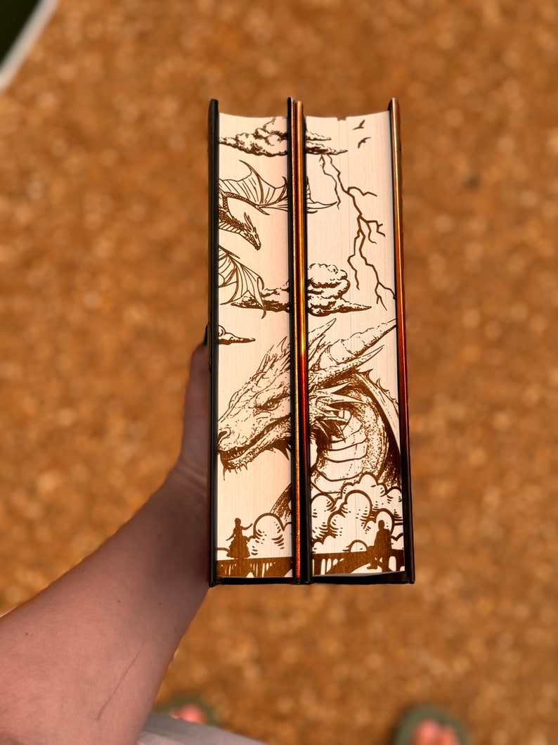 May include: Two hardback books with cream-coloured covers featuring a dragon illustration. The dragon is depicted flying through clouds with lightning bolts in the background. The books have black spines and gold accents.