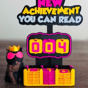 May include: A 3D-printed display featuring a black sign with the text NEW ACHIEVEMENT YOU CAN READ in pink and yellow. Below is a counter showing 004 with stacks of yellow and pink tiles. A black dog figurine wears a yellow crown and pink glasses.