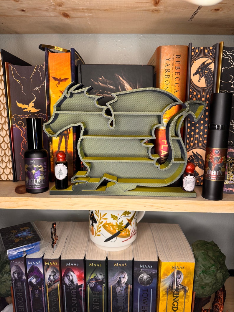 May include: A green dragon-shaped bookshelf with four shelves. The bookshelf is on a wooden shelf with a variety of books, a mug, and bottles. The books include titles like "Tower of Dawn" and "Queen of Shadows".