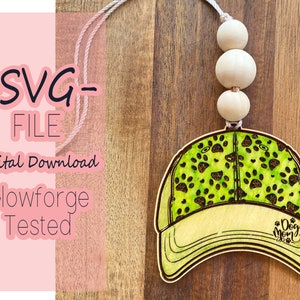 May include: A wooden cutout of a baseball cap with a green paw print pattern and the text "Dog Mom" on the front. The cap is attached to a string with wooden beads.