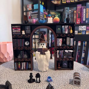 Puede incluir: Una figurita de fantasma blanco leyendo un libro titulado "Harry Potter" en un escenario de estantería en miniatura. La estantería está llena de libros y tiene un arco negro. Hay velas, jarrones y otros artículos decorativos en los estantes.