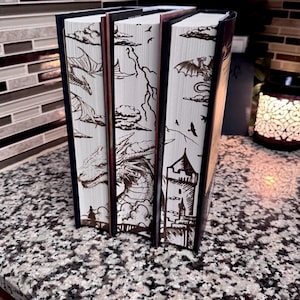 May include: Three hardback books with white covers and brown illustrations of dragons, clouds, and a castle. The books are arranged vertically, with the tallest book on the right.