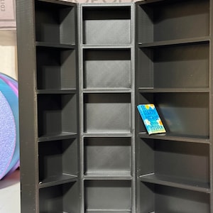May include: Black, three-sectioned, open shelf display with a small blue book titled "It Starts With Us" by Colleen Hoover on the middle shelf.