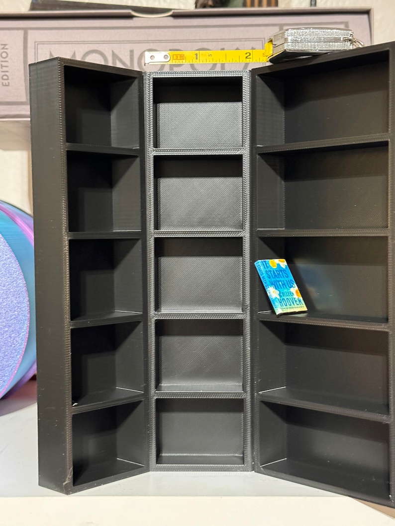 May include: Black, three-sectioned, open shelf display with a small blue book titled "It Starts With Us" by Colleen Hoover on the middle shelf.