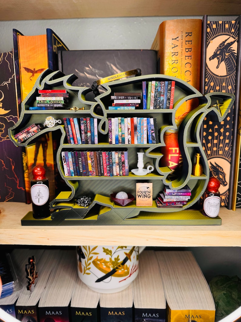 May include: A green dragon-shaped bookshelf with multiple shelves filled with books. The bookshelf is on a wooden shelf with a mug and more books below it. The bookshelf has a gold bracelet, a silver candlestick, and a small book titled "Fourth Wing" on it.