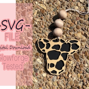 May include: A wooden cutout of a giraffe head with a black and brown spotted pattern. The cutout is attached to a string with wooden beads. The text "SVG FILE Digital Download Glowforge Tested" is on a pink background.