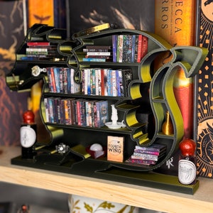 May include: A green and gold dragon-shaped bookshelf with books on the shelves. The bookshelf is on a wooden shelf with a mug and other decorative items.