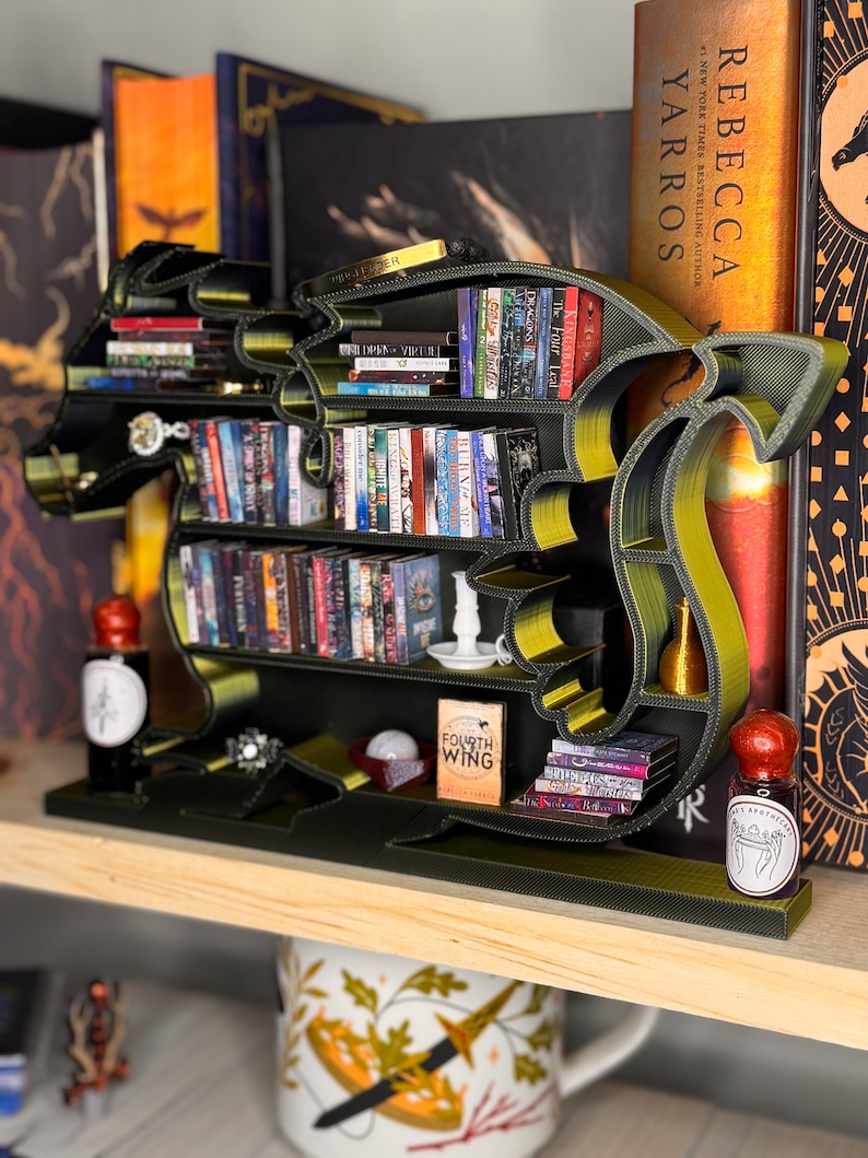 May include: A green and gold dragon-shaped bookshelf with books on the shelves. The bookshelf is on a wooden shelf with a mug and other decorative items.
