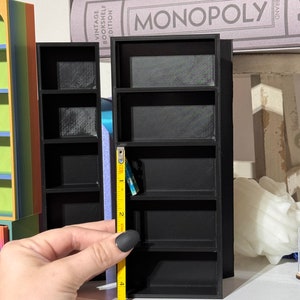 May include: Two black miniature bookshelves with four shelves each. The shelves are made of 3D printed plastic. A yellow measuring tape is next to the bookshelf on the right, measuring 5 inches.