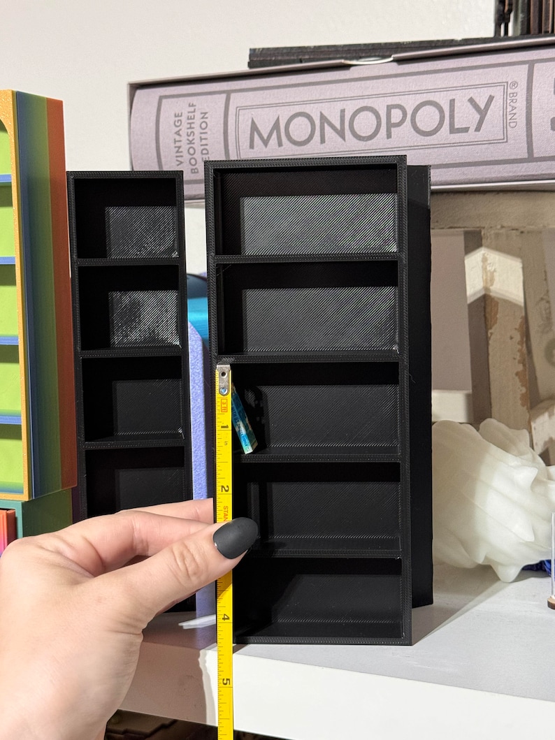 May include: Two black miniature bookshelves with four shelves each. The shelves are made of 3D printed plastic. A yellow measuring tape is next to the bookshelf on the right, measuring 5 inches.
