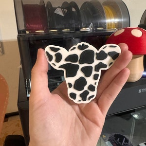 May include: A 3D printed cow ornament with a black and white spotted pattern. The ornament is made of white plastic and has a smooth, glossy finish.