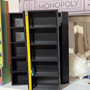 May include: A black miniature bookshelf with six shelves, measuring 7 inches tall. The bookshelf is on a white surface with a yellow measuring tape next to it.