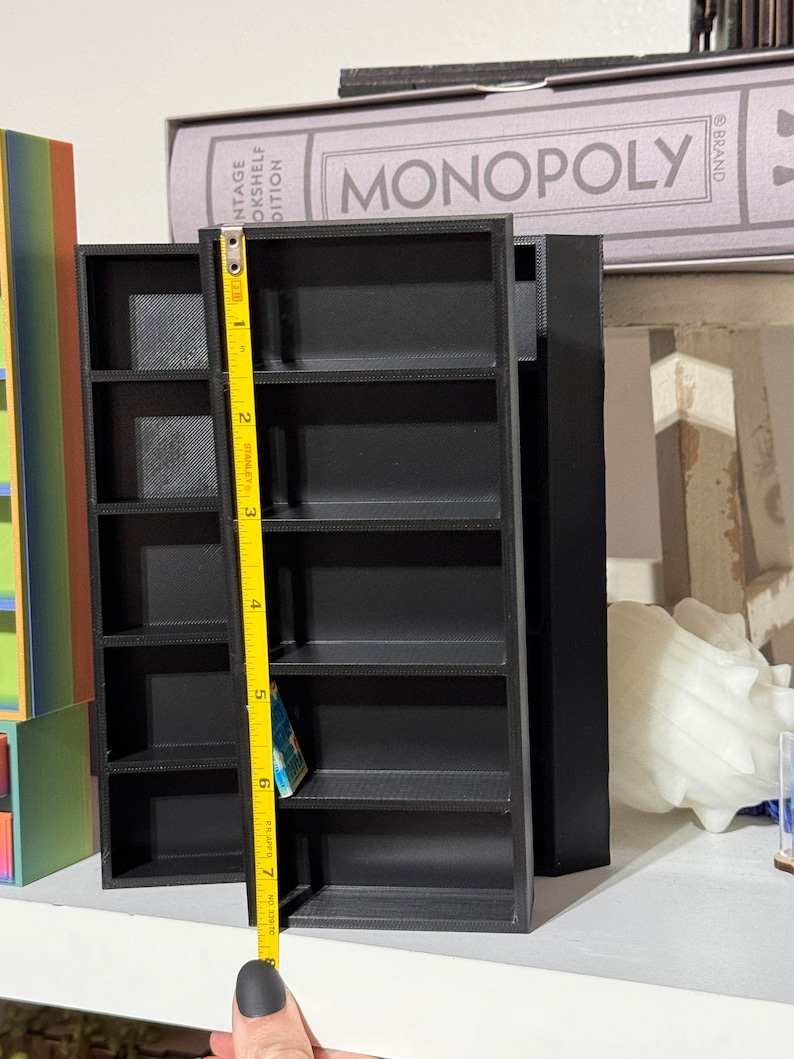 May include: A black miniature bookshelf with six shelves, measuring 7 inches tall. The bookshelf is on a white surface with a yellow measuring tape next to it.