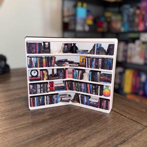 May include: A white bookshelf with multiple shelves filled with colorful books. The bookshelf is in a corner and has a decorative skull on the top shelf.