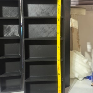 May include: Black plastic shelf unit with five shelves, measuring 8 inches tall. The shelf unit is standing on a table.