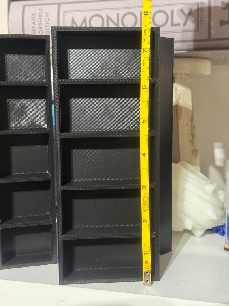 May include: Black plastic shelf unit with five shelves, measuring 8 inches tall. The shelf unit is standing on a table.