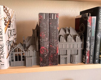 May include: A gray 3D printed house-shaped bookend with two books titled "Hunting Adeline" and "Haunting Adeline" by H.D. Carlton. The bookend is on a wooden shelf with other books.