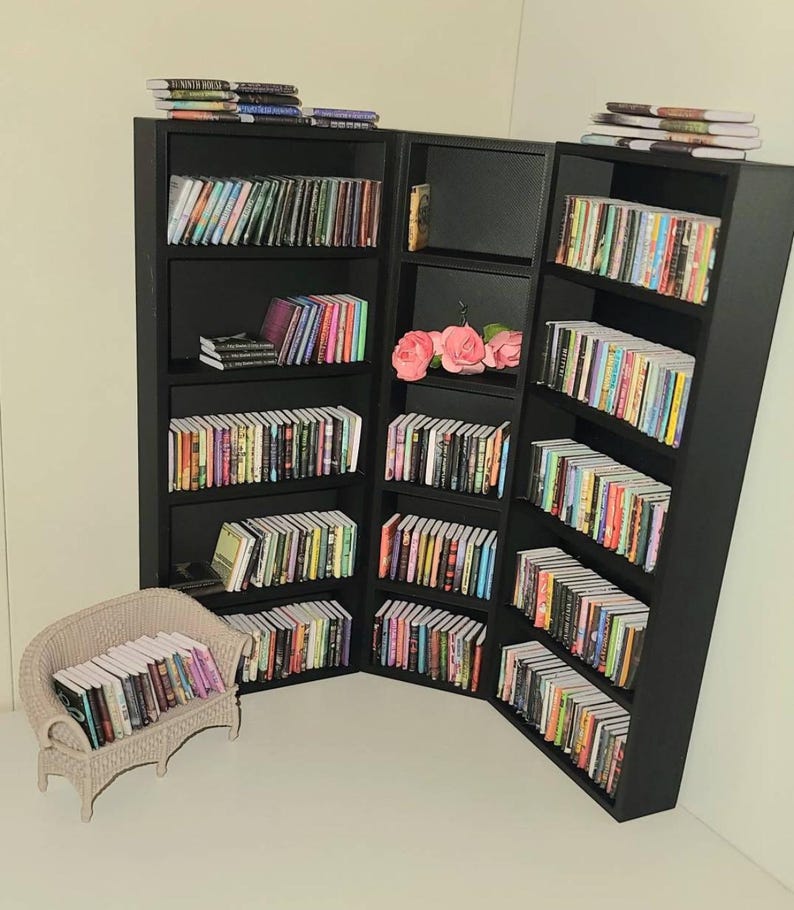May include: A black bookshelf with multiple shelves filled with colorful books. The bookshelf is in a corner of a room and has a small white wicker chair with books on it.