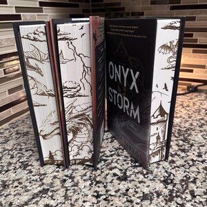 May include: A set of five fantasy novels with black spines and white covers featuring dragon and castle illustrations. The title of the centre book is "ONYX STORM" by Rebecca Yarros.