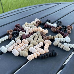 Könnte beinhalten: Eine Sammlung bunter knochenförmiger Perlen in verschiedenen Brauntönen, Beige und Weiß. Die Perlen liegen lose auf einer dunkelgrauen Oberfläche.