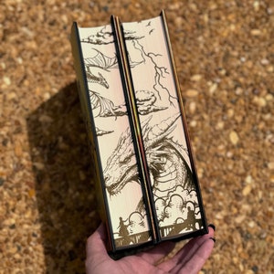 May include: Two hardback books with black spines and gold accents. The covers feature a detailed illustration of a dragon with clouds and lightning in the background.