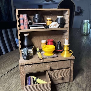 May include: A miniature brown wooden cabinet with shelves and drawers, filled with various items. Includes books, a skull, potions, a mortar and pestle, a candle holder, and a small cactus. The word "GRIMOIRE" is visible on a book.