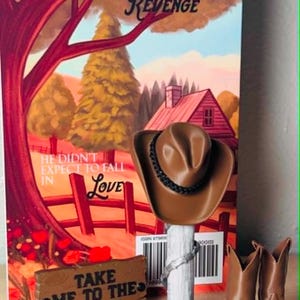 Puede incluir: Un sombrero de vaquero marrón sobre un soporte de poste blanco, un par de botas de vaquero marrones y un letrero rectangular marrón con el texto "TAKE ME TO THE RANCH" en letras negras frente a una portada de libro con un árbol rojo y una casa.