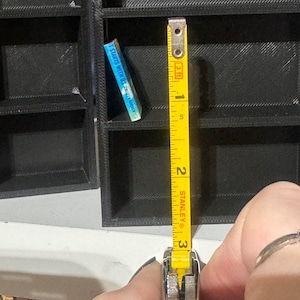 May include: A yellow tape measure with black markings is extended to show 3 inches. The tape measure is held in a hand. The tape measure is in front of a black bookshelf with a small blue book titled "It Starts With Us" by Colleen Hoover.