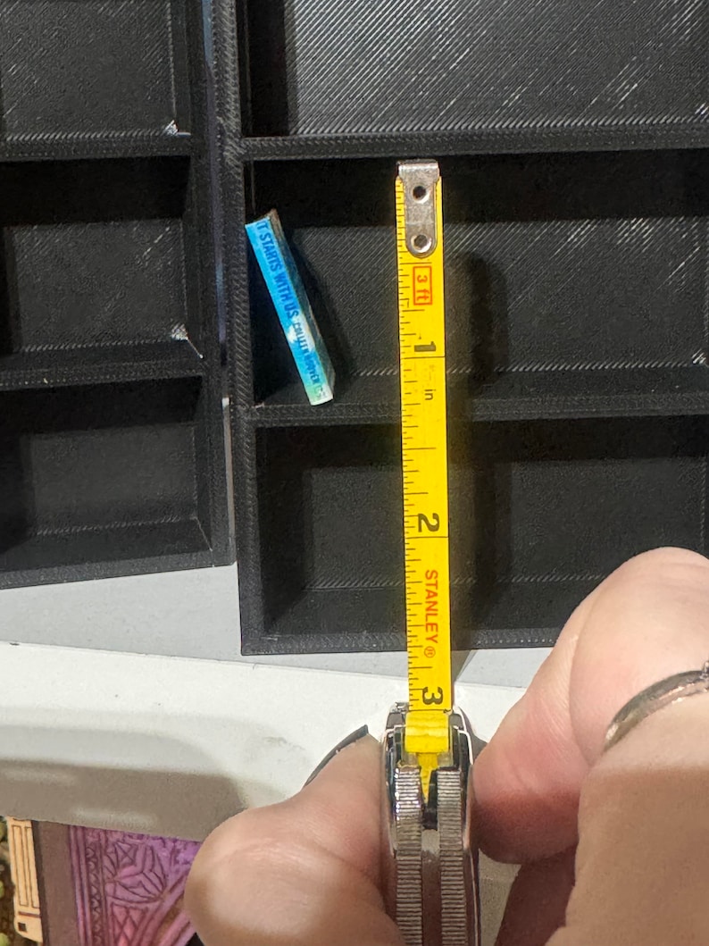 May include: A yellow tape measure with black markings is extended to show 3 inches. The tape measure is held in a hand. The tape measure is in front of a black bookshelf with a small blue book titled "It Starts With Us" by Colleen Hoover.
