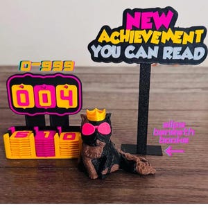 May include: A 3D-printed black and brown cat figurine wearing a yellow crown and pink sunglasses, accompanied by a black sign reading NEW ACHIEVEMENT YOU CAN READ and a black counter display with yellow and pink tiles showing 0-999 and slips beneath books.