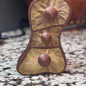 May include: A brown, three-drawer jewelry box with a unique, curved shape. Each drawer front features a radial design in shades of gold and brown, with a circular knob. The box is made of wood and sits on a speckled surface.