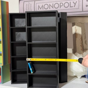 May include: A black miniature bookshelf with five shelves, measuring 5 inches in length. A small blue book is on the third shelf. The bookshelf is on a white surface.