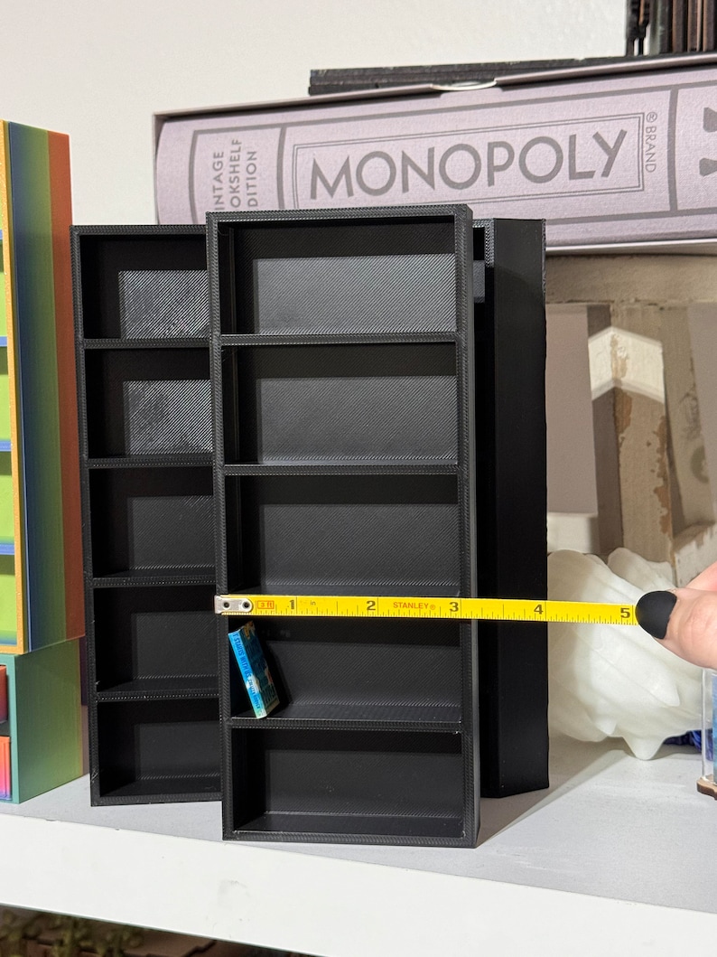 May include: A black miniature bookshelf with five shelves, measuring 5 inches in length. A small blue book is on the third shelf. The bookshelf is on a white surface.