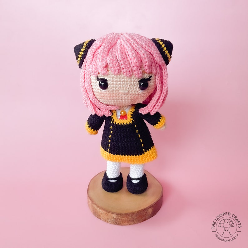 May include: A pink and black crocheted doll with black eyes and a black and yellow dress. The doll is standing on a wooden base.
