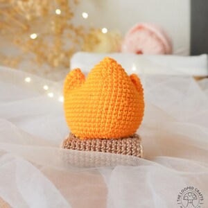 May include: A crocheted campfire with a bright orange flame and a brown base. The campfire is sitting on a white fabric surface.