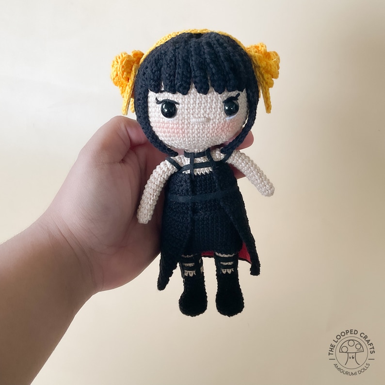 May include: A crocheted doll with black hair, a black dress, and yellow hair bows. The doll is holding a red and black cape.
