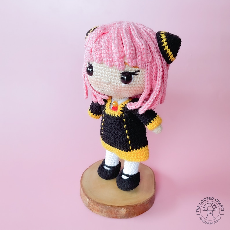 May include: A pink and black crocheted doll with yellow accents. The doll is wearing a black dress with yellow trim and has a pink wig. The doll is standing on a wooden base.