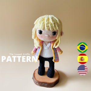 Puede incluir: Muñeca amigurumi de crochet con cabello rubio, vistiendo una chaqueta estampada en rosa y azul y pantalones negros. La muñeca está de pie sobre una base de madera. La palabra "PATTERN" está escrita en letras grandes en el fondo.