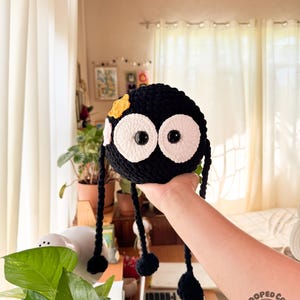 May include: A round, black crocheted plush toy featuring two large white circular eyes with black pupils, a small yellow star-shaped accent, and four long, thin black dangling legs with rounded ends. The plush has a soft, textured yarn finish.