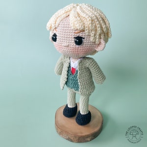 May include: A crocheted doll with blonde hair wearing a green jacket and black shoes. The doll is standing on a wooden base.