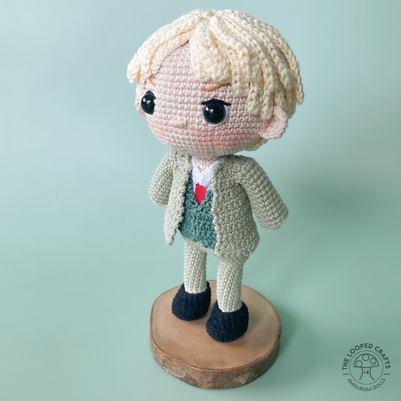 May include: A crocheted doll with blonde hair wearing a green jacket and black shoes. The doll is standing on a wooden base.