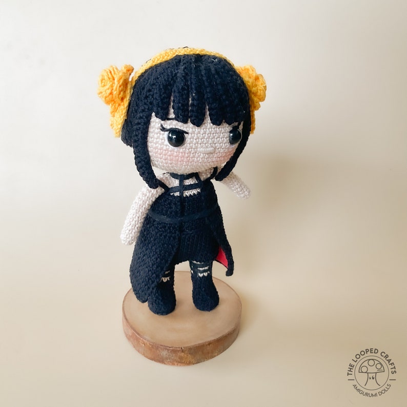 May include: A crocheted doll with black hair, yellow hair bows, and a black dress. The doll is standing on a wooden base.