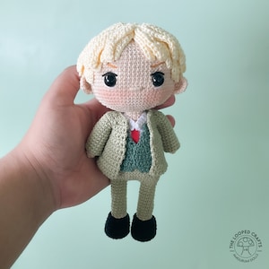 May include: A crocheted amigurumi doll with blonde hair, wearing a green jacket and a red tie. The doll is holding a small red heart in its hand.