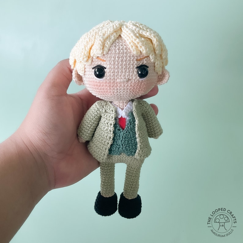 May include: A crocheted amigurumi doll with blonde hair, wearing a green jacket and a red tie. The doll is holding a small red heart in its hand.