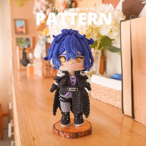 May include: A crocheted doll with blue hair and amber eyes, wearing a black coat with silver accents over grey pants. The doll stands on a circular wooden base. Text at the top reads The Looped Crafts PATTERN in white capital letters.