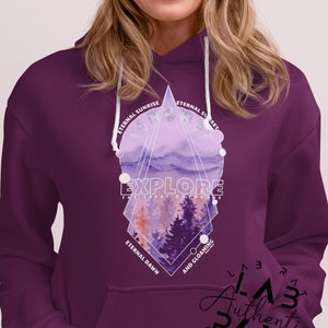 May include: Purple hoodie with a geometric design featuring a watercolor landscape with trees and the text "EXPLORE THE GREAT OUTDOORS" in white. The design also includes the text "ETERNAL SUNRISE", "ETERNAL SUNSET", "ETERNAL DAWN", and "AND GLOAMING". The hoodie has a white drawstring and a kangaroo pocket.