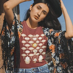 May include: A woman wearing a maroon t-shirt with a graphic print of eyes and hands, a floral print kimono, denim shorts, and a black hat. The t-shirt has the text "Authentic" printed on it.