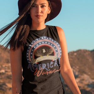 May include: A woman wearing a black sleeveless shirt with a graphic of an eagle and stars with the text "American Woman". She is wearing a brown hat and denim shorts. The background is a mountain range.