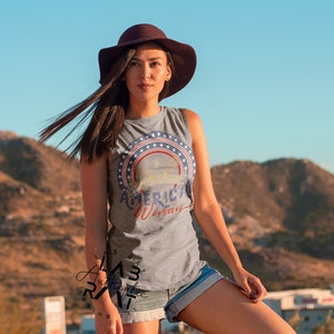 May include: A woman wearing a grey sleeveless shirt with a red, white, and blue graphic that says "American Woman". She is wearing a brown hat and denim shorts. The background is a blurry image of a mountain.